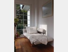 Helles Wohnzimmer mit einer weißen Liege auf einem Holzboden, daneben ein großes Fenster mit Sprossenrahmen, das den Blick auf grüne Bäume freigibt. An der Wand hängt ein minimalistisches Bild, und eine Zimmerpflanze steht im Vordergrund.