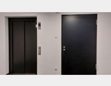 Schlichte Innenwand mit einem geschlossenen schwarzen Aufzug auf der linken Seite und einer schwarzen Zimmertür rechts, umgeben von weiß gestrichenen Wänden. Ein leuchtendes Display über dem Aufzug zeigt die Zahl 1.