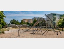 Spielplatz mit einem großen Klettergerüst aus Stahlstangen und Netzseilen sowie einer schlangenförmigen Schaukel, umgeben von sandigem Untergrund. Im Hintergrund sind Wohngebäude, Bäume und eine kleine Wiese zu sehen.