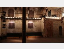 Industrieller Innenraum mit rauer, unverputzter Stein- und Ziegelwand. Eine schmale hölzerne Galerie mit Lichterketten verläuft entlang der oberen Wand, unten befinden sich eine Sitzbank und eine große Holztür.