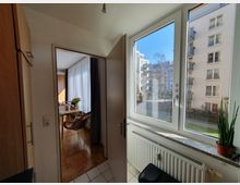 Blick aus einer kleinen Innenraumküche mit Fliesenboden durch ein Fenster auf ein Wohngebäude mit Balkonen. Im angrenzenden Raum mit Parkettboden stehen ein Tisch, Stühle und Blumen in einer Vase; Tageslicht fällt durch weiße Vorhänge.