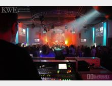 Innenraum eines Veranstaltungszentrums mit einer Bühne im Hintergrund, auf der eine Band unter bunten Scheinwerferlichtern auftritt. Im Vordergrund ist ein Mischpult zu sehen, dahinter stehen viele Zuschauer in einem beleuchteten Raum mit Werbebildschirmen an den Wänden.