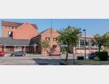 Backstein-Gebäudeensemble mit einer Mischung aus flachen und steilen Dächern, links ein größeres, fensterarmes Gebäude, rechts eine verglaste Halle mit der Aufschrift „Theater Café“. Vor dem Gebäude befinden sich geparkte Autos und vereinzelte Bäume an einer asphaltierten Straße.