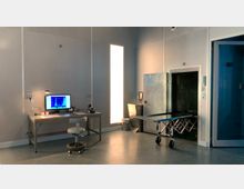 Ein moderner Raum mit klinischer Ausstattung, darunter ein Metalltisch auf Rollen, ein Schreibtisch mit Monitor und Arbeitsmaterialien, sowie eine helle Wandbeleuchtung. Rechts ist eine Kühlkammer mit Edelstahlklapptür zu sehen.