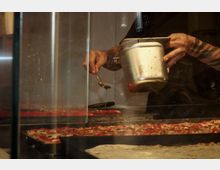 Ein Koch steht hinter einer Glasscheibe und verteilt mit einem Löffel flüssiges Öl oder eine andere Zutat aus einem Metallbehälter auf rechteckige Pizzen, die auf großen Backblechen liegen. Im Vordergrund sind verschiedene Pizzen mit unterschiedlichen Belägen zu sehen.