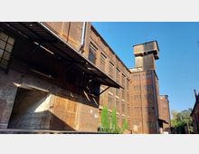 Backstein-Industriegebäude mit großen Fenstern und einem hohen Turm, umgeben von einem offenen Hof. Der Eingang einer Lagerhalle ist im Vordergrund und weist metallene Überdachungen sowie verwitterte Wände auf; im Hintergrund sind Bäume und blauer Himmel sichtbar.