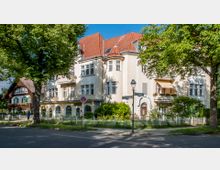 Mehrfamilienhaus mit heller Fassade und roten Dachziegeln in einer baumbestandenen Wohnstraße. Vor dem Gebäude befinden sich ein kleiner Garten mit Zaun sowie ein Altstadtlaternenpfahl, daneben ein Hinweisschild der "Teutonenstraße". Im Hintergrund ist ein Haus im Landhausstil mit Holzdetails und Fensterläden zu sehen.