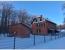 Zwei Gebäude aus rotem Backstein, eines mit Fachwerkelementen, stehen in einer verschneiten Landschaft, umgeben von einem Metallzaun und kahlen Bäumen. Der Himmel ist klar und blau.