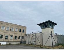 Außenansicht eines umzäunten Gefängnisareals mit hohem Maschendrahtzaun und Stacheldraht, daneben ein Wachtturm mit verglaster Aussichtsplattform. Links steht ein mehrstöckiges, rechteckiges Gebäude mit vielen Fenstern unter bewölktem Himmel.