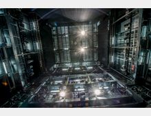 Blick in den Bühnenraum eines modernen Theaters nach oben, mit mehreren Scheinwerfern, Metallgittern und technischen Geräten an schwarzen Gerüststrukturen. Die Umgebung ist dunkel, mit starker künstlicher Beleuchtung.