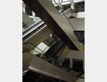 Mehrstöckiger Innenraum mit mehreren modernen Rolltreppen, die sich kreuzen, um verschiedene Ebenen des Gebäudes zu verbinden. Die Umgebung ist durch eine weiße Wandgestaltung und klare Linien geprägt.