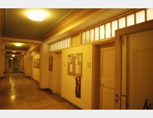 Flur in einem Gebäude mit gelblichen Wänden und Deckenbeleuchtung. Entlang der Wände befinden sich geschlossene Türen, Pinnwände mit Aushängen und oberhalb der Türen sind Fenster eingesetzt.