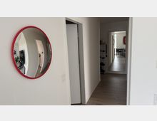 Heller Wohnungsflur mit weißen Wänden und dunkelgrauem Holzfußboden; links hängt ein runder Spiegel mit rotem Rahmen an der Wand. Am Ende des Gangs führt eine offene Tür in einen weiteren Raum, daneben steht ein schmales Regal.