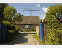 Einfahrt zu einem ländlichen Grundstück mit einer alten Scheune im Hintergrund, deren Dach mit roten Ziegeln gedeckt ist. Seitlich stehen Bäume und Sträucher, ein blauer Torflügel befindet sich rechts im Vordergrund, und im Hintergrund sind Hochspannungsleitungen sichtbar.