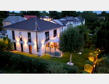 Modernes zweistöckiges Wohnhaus mit weißer Fassade und dunklem Walmdach, beleuchtet von Außenlampen in der Dämmerung. Das Haus ist umgeben von einem gepflegten Garten mit Rasen, Bäumen und Hecken, teils ebenfalls durch Gartenlichter beleuchtet.