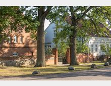 Backsteinbau eines historischen Gebäudes mit Rundbogenfenstern, daneben ein helles, stuckverziertes Haus. Im Vordergrund stehen zwei große Bäume, und eine Ziegelmauer mit einem Eisentor markiert den Eingangsbereich.