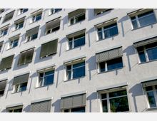 Weiße Fassade eines mehrstöckigen Gebäudes mit mehreren rechteckigen Fenstern, von denen einige mit Rollläden teilweise oder vollständig abgedeckt sind. Die Fensterrahmen sind weiß, und die Schatten der Rollläden zeichnen sich auf der Fassade ab.