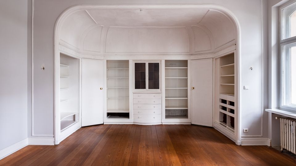 Leerer Innenraum mit hoher Decke und großem, bogenförmigem Wandeinbau aus weißen Einbauschränken und Regalen, inklusive Schubladen in der Mitte. Davor liegt ein dunkler Holzdielenboden; rechts befindet sich ein großes Fenster mit Heizkörper darunter.