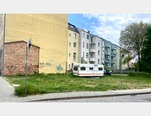 Eine kleine Grünfläche mit hohem Gras, auf der ein weißer Wohnwagen parkt. Im Hintergrund befinden sich mehrstöckige Wohnhäuser, eines davon mit sichtbaren Graffiti an einer gelben Fassadenwand, sowie ein großer Baum am rechten Bildrand.