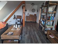 Wohnzimmer mit Dachschrägen, Holzbalken und Holzboden. Der Raum enthält Möbel im rustikalen Stil, darunter ein Couchtisch, ein Bücherregal, eine Couch und eine Kommode; links steht eine alte Nähmaschine als Dekoration, im Hintergrund eine Wanduhr.