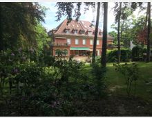 Zweigeschossiges rotes Wohnhaus mit Satteldach und mehreren Dachfenstern, umgeben von einem weitläufigen Garten mit Bäumen, Blumen und Sträuchern. Im Vordergrund sind blühende Rhododendren und eine Wiese zu sehen.