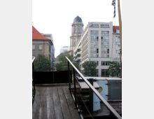 Holzsteg eines Bootes im Vordergrund, der zu einer Stadtansicht mit modernen Bürogebäuden und einem historischen Turm mit Kuppel im Hintergrund führt. Bäume säumen den Straßenbereich zwischen den Gebäuden.