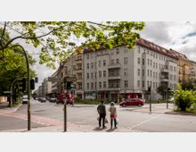 Straßenszene mit Kreuzung in einer urbanen Umgebung, flankiert von mehrstöckigen Wohnhäusern mit hellen Fassaden und roten Ziegeldächern. Im Vordergrund stehen zwei Personen an einer Ampel, umgeben von Straßenschildern, Bäumen und parkenden Autos.