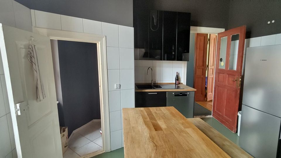 Moderne Küche mit weißen Wandfliesen und dunkelgrauen Wänden, schwarzer Hängeschrankzeile, Spüle und integrierter Spülmaschine sowie großem Holztisch im Vordergrund. Rechts stehen ein hoher Kühlschrank und ein Backofen; offene Türen führen in einen Flur und einen angrenzenden Raum.