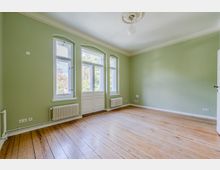 Heller, leerer Wohnraum mit hellgrünen Wänden, hohen Decken mit Stuckrosette und breiten Holzdielen. Drei hohe Fenster und eine verglaste Flügeltür lassen Tageslicht herein; unter den Fenstern stehen weiße Heizkörper.