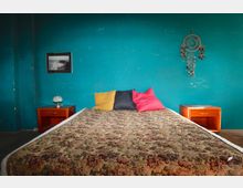 Schlafzimmer mit türkisfarbener Wand, einem Bett mit floral gemusterter Tagesdecke und farbigen Kissen. Rechts und links vom Bett stehen Nachttische aus Holz; an der Wand hängen ein gerahmtes Bild und ein Traumfänger.