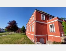 Rotes zweistöckiges Gebäude mit weißen Fenstern und Fassadendetails, umgeben von einer Rasenfläche mit Bäumen; im Hintergrund weitere Gebäude und ein blauer Himmel.