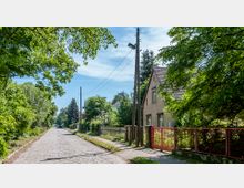 Eine gepflasterte, baumbestandene Dorfstraße mit Strommasten und einem Fußweg auf der rechten Seite. Rechts steht ein Haus mit rotem Ziegeldach, einem kleinen Garten und einem Zaun mit rotem Tor.
