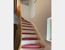 Eine geschwungene Innentreppe aus hellem Holz mit roten Teppichläufern auf jeder Stufe. Die Treppe ist von weißen Wänden umgeben, und rechts gibt es ein kleines Fenster mit einer hölzernen Fensterbank.