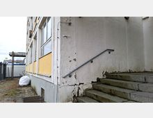 Außenansicht eines älteren Mehrgeschossgebäudes mit großen Fenstern und abblätterndem Putz an der Fassade. Rechts führt eine steinerne Treppe mit Metallhandlauf zu einem erhöhten Eingang, daneben ein gepflasterter Weg entlang der Gebäudeseite.