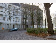 Kopfsteinpflasterstraße in einem Wohngebiet mit mehrstöckigen Gebäuden im Altbaustil, teils in grau und blau gestrichen. Im Vordergrund stehen Bäume mit kahlen Zweigen und ein kleiner begrünt und mit Herbstlaub bedeckter Platz, während auf der Straße geparkte Autos und ein Fahrrad zu sehen sind.