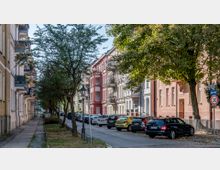 Straße mit mehrstöckigen Altbauten auf beiden Seiten, verziert mit Balkonen und bunten Fassaden; schmale Gehwege, parkende Autos entlang der Straße, Bäume und eine Laterne im Vordergrund, sowie ein Geschwindigkeitsbegrenzungsschild.