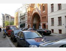 Straßenszene in einer Stadt mit geparkten Autos und Motorrad am rechten Straßenrand. Auf der rechten Seite befindet sich ein historisches Backsteingebäude mit einem großen, torbogenförmigen Eingang, flankiert von Wohnhäusern, und im Hintergrund ist ein größeres Gebäude mit gotischen Elementen sichtbar.