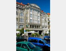 Historisches Gebäude mit klassizistischer Fassade, hohen weißen Fenstern und dekorativen Elementen, darunter ein halbrundes Giebelfeld mit Relief. Vor dem Gebäude befinden sich ein Café mit Sonnenschirmen, parkende Autos und Vegetation.