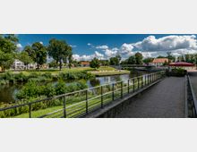 Flusslandschaft in einer städtischen Umgebung mit Gehwegen, Geländern und einem parkähnlichen Uferbereich. Im Hintergrund sind eine Brücke, Bäume und einzelne Gebäude sowie ein Sonnenschirm eines Cafés sichtbar.