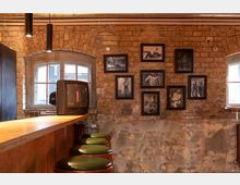 Ziegelsteinwand mit zwei Fenstern und einer Sammlung schwarz-weißer Fotografien in Bilderrahmen. Im Vordergrund eine Holztheke mit alten Barhockern und ein Röhrenfernseher.