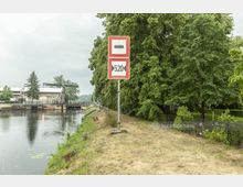 Ein Wasserkanal mit einer begrünten Uferböschung und einem Verkehrsschild, das eine Höhenbegrenzung von 5,20 Metern anzeigt. Im Hintergrund befindet sich eine Klappbrücke und ein Gebäude, umgeben von großen Bäumen.
