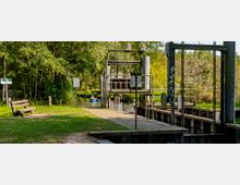 Eine kleine Schleusenanlage an einem schmalen Fluss, umgeben von üppigem Grün und Bäumen. Im Hintergrund paddeln Personen in einem blauen Kajak, rechts und links sind Holzstege und Metallkonstruktionen der Schleuse zu sehen, daneben eine Sitzbank und ein Wegweiser.