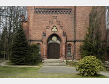 Backsteinfassade einer Kirche mit mittelalterlich inspiriertem Eingangsportal, verziert mit Glasfenstern und kleinen Ornamenten. Vor dem Gebäude führen Stufen zum Holztor, umgeben von Bäumen und Sträuchern in einer parkähnlichen Umgebung.