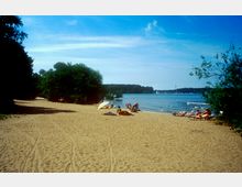 Sandstrand mit vereinzelten Badegästen, die auf Handtüchern oder Liegen entspannen, umgeben von Bäumen und Büschen. Im Hintergrund ist ein ruhiger See mit leichtem Wellengang und einem bewaldeten Ufer zu sehen.