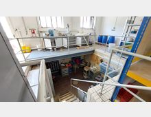 Mehrstöckige Werkstatt mit einem oberen Bereich, der durch Metallgeländer geschützt ist, und Arbeitsflächen mit Laborgeräten und blauen Fässern. Im unteren Bereich befinden sich Werkbänke, Werkzeuge, Lagerregale und eine Fliesentreppe, die die Ebenen verbindet.