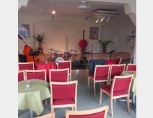 Kleiner Veranstaltungsraum mit einer Bühne im Hintergrund, auf der ein Schlagzeug, ein Kontrabass und weitere Musikinstrumente stehen. Im Vordergrund befinden sich Stuhlreihen mit roten Polstern und runde Tische mit farbigen Tischdecken.