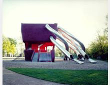 Spielplatz mit einer auffälligen, modernen Kletter- und Rutschstruktur: Ein rot-lila Gebäude mit mehreren silbernen Rutschen, Kletternetzen und Treppen, umgeben von Sandboden, einer Wiese und Bäumen im Hintergrund.