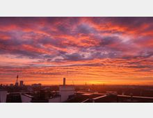 Städtische Skyline mit Blick auf Dächer im Vordergrund und dem Fernsehturm im Hintergrund, vor einem dramatischen Himmel in kräftigen Orange- und Lilatönen während eines Sonnenuntergangs.