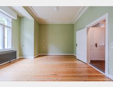 Leerer Wohnraum mit hellgrünen Wänden, hohen Fenstern und einem Heizkörper unter dem Fenster sowie breiten Holzdielenboden. Stuckverzierte Deckenleisten und eine weiße Tür führen in einen angrenzenden Flur mit weiteren Türen.