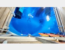 Blick von oben auf ein großes, tiefes Schwimmbecken mit blauem Wasser, in dem Taucher erkennbar sind. Der Rand des Beckens ist von Plattformen aus Holz und Metallgeländern umgeben, mit Rettungsausrüstung an der Seite.